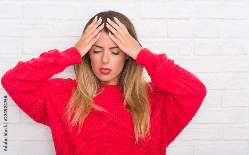 Young adult woman over white brick wall wearing winter outfit at home suffering from headache desperate and stressed because pain and migraine. Hands on head. Young adult woman over white brick wall wearing winter outfit at home suffering from headache desperate and stressed because pain and migraine. Hands on head.