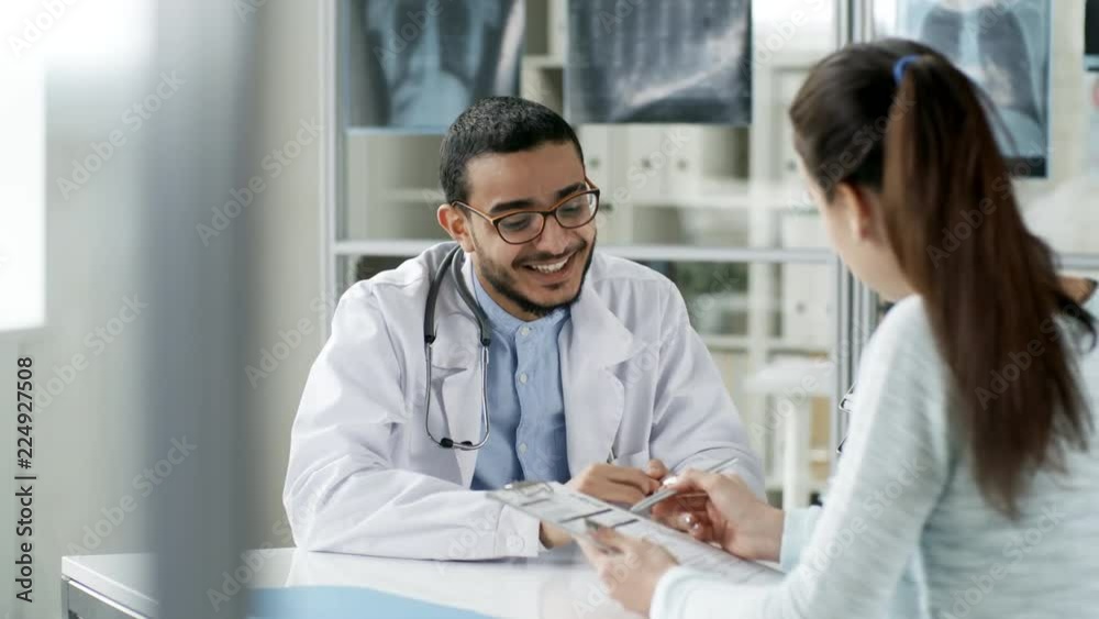 Tracking shot of smiling Asian doctor in lab coat and glasses sitting at desk in office and talking to female patient who is filling out form with information Tracking shot of smiling Asian doctor in lab coat and glasses sitting at desk in office and talking to female patient who is filling out form with information