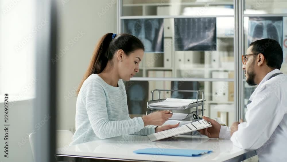 Tracking shot of young Caucasian woman filling out form and talking to Asian doctor before health checkup, side view Tracking shot of young Caucasian woman filling out form and talking to Asian doctor before health checkup, side view