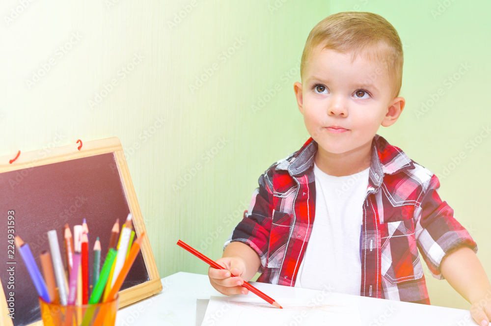 Cute little boy in a red checkered shirt dreams and draws with a red pencil in a white album Cute little boy in a red checkered shirt dreams and draws with a red pencil in a white album