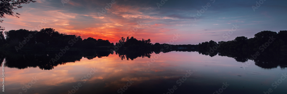 Reflection of a lake creating perfect symmetry with abstract shapes of the forest tree silhouettes. At colorful red and orange sunset sky. Braunschweig, Germany