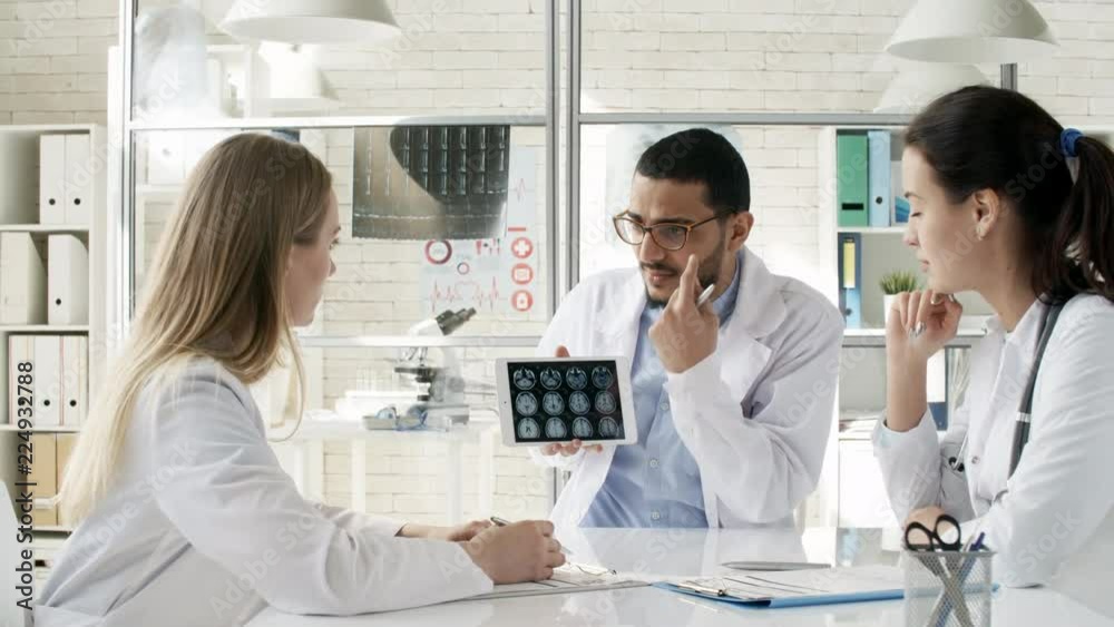Zoom out shot of friendly black man in lab coat using tablet computer to show MRI images and explaining them to two female colleagues during meeting in clinic