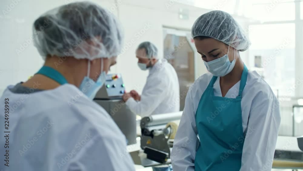 Three colleagues in protective clothing, aprons, bouffant mob caps, and face masks working in food production line at factory Three colleagues in protective clothing, aprons, bouffant mob caps, and face masks working in food production line at factory