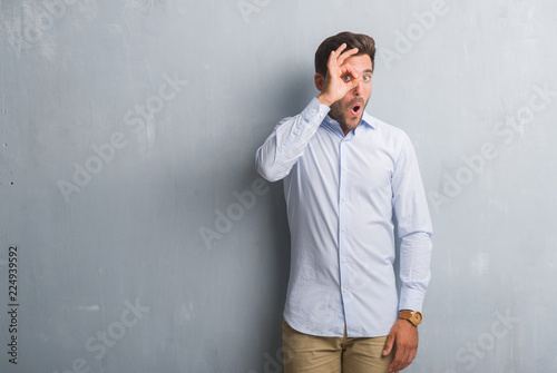 Handsome young business man over grey grunge wall wearing elegant shirt doing ok gesture shocked with surprised face, eye looking through fingers. Unbelieving expression.