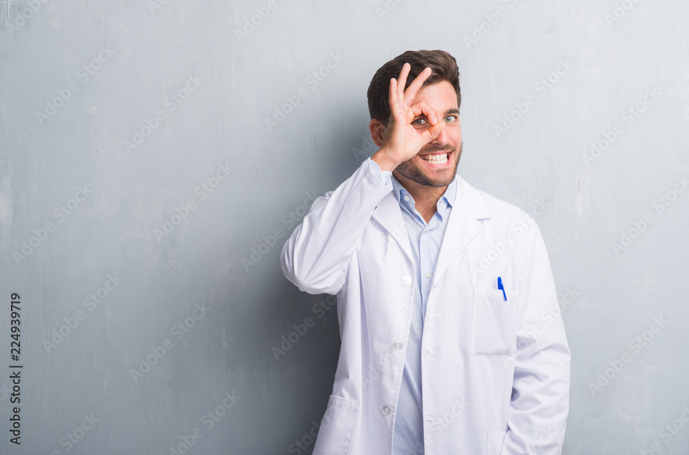 Handsome young professional man over grey grunge wall wearing white coat doing ok gesture with hand smiling, eye looking through fingers with happy face. Handsome young professional man over grey grunge wall wearing white coat doing ok gesture with hand smiling, eye looking through fingers with happy face.
