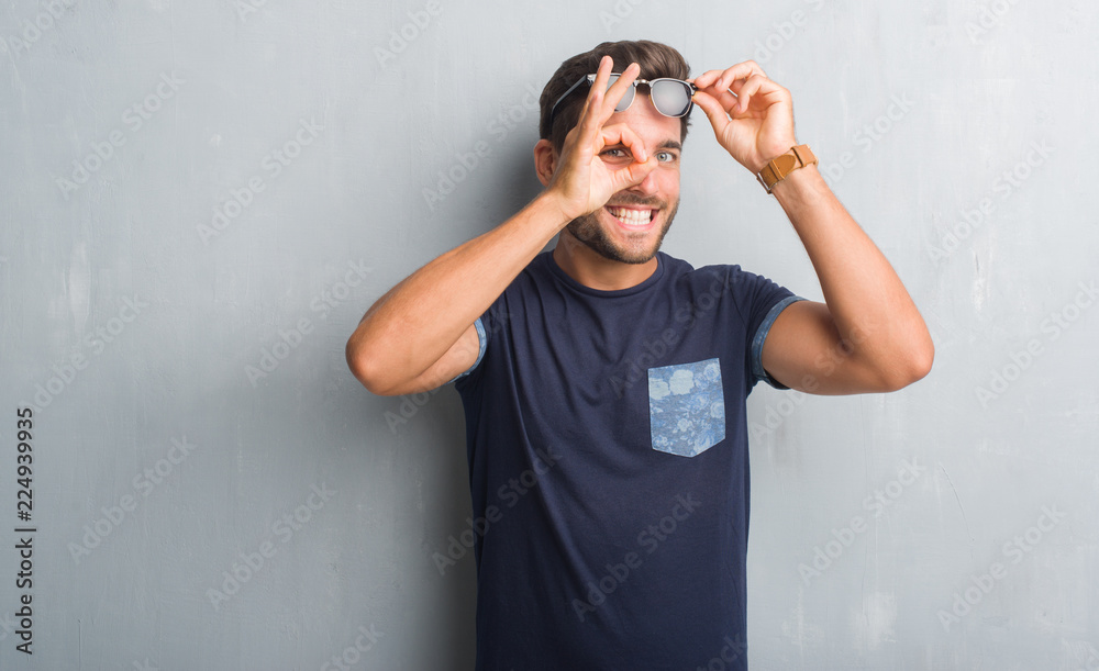 Naklejka premium Handsome young man over grey grunge wall wearing sunglasses doing ok gesture with hand smiling, eye looking through fingers with happy face.