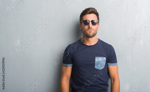 Handsome young man over grey grunge wall wearing sunglasses with serious expression on face. Simple and natural looking at the camera.