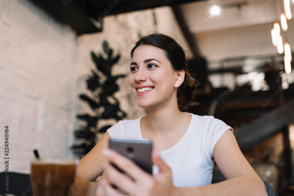 Young attractive woman with beautiful face and smile holding mobile phone, downloading application in loft cafe. Social media marketing. Female blogger write blog post using smartphone and internet. Young attractive woman with beautiful face and smile holding mobile phone, downloading application in loft cafe. Social media marketing. Female blogger write blog post using smartphone and internet.