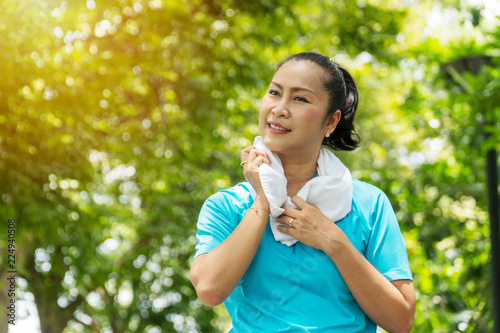 Elderly lifestyle concept. Attractive senior women 50s smiling while wiping her throat with white towel on neck after exercises in the public park.