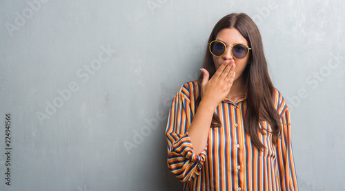 Young brunette woman over grunge grey wall wearing retro sunglasses and outfit cover mouth with hand shocked with shame for mistake, expression of fear, scared in silence, secret concept