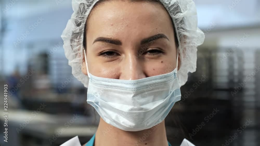 Close up face of young female doctor or factory worker wearing bouffant mob cap and face mask turning at camera and smiling, tracking right