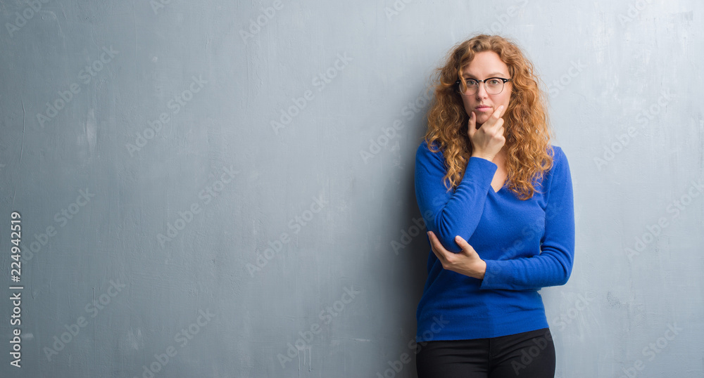 Fototapeta premium Young redhead woman over grey grunge wall looking confident at the camera with smile with crossed arms and hand raised on chin. Thinking positive.