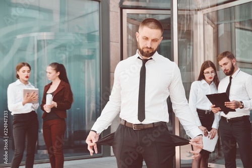 Bankrupt. A young man in black trousers and a white shirt demonstrates his empty pockets in the background of a group of office workers. Dismissal from work.