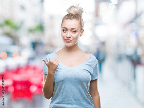 Young blonde woman over isolated background smiling with happy face looking and pointing to the side with thumb up.