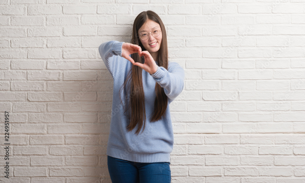 Young Chinise woman over white brick wall smiling in love showing heart symbol and shape with hands. Romantic concept.