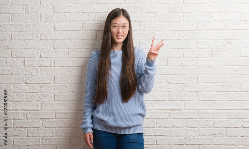 Young Chinise woman over white brick wall showing and pointing up with fingers number three while smiling confident and happy. Young Chinise woman over white brick wall showing and pointing up with fingers number three while smiling confident and happy.