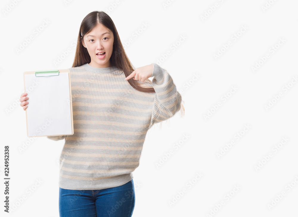 Young Chinese woman over isolated background holding clipboard with surprise face pointing finger to himself Young Chinese woman over isolated background holding clipboard with surprise face pointing finger to himself
