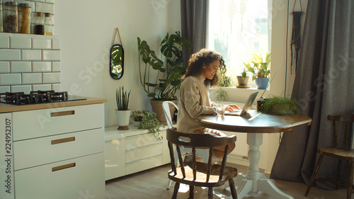 Charming young woman typing on laptop computer in a kitchen.
