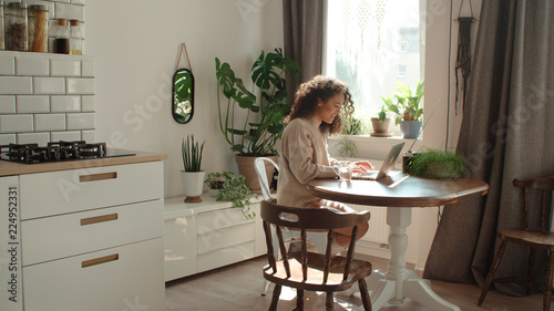 Charming young woman typing on laptop computer in a kitchen.