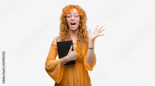 Young redhead student woman holding a book very happy and excited  winner expression celebrating victory screaming with big smile and raised hands