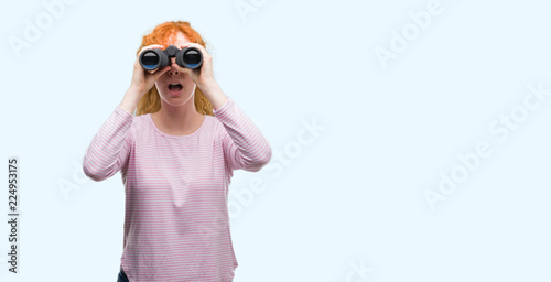 Young redhead woman looking through binoculars scared in shock with a surprise face, afraid and excited with fear expression © Krakenimages.com