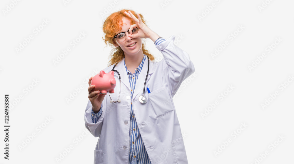 Young redhead doctor woman holding piggy bank with happy face smiling doing ok sign with hand on eye looking through fingers Young redhead doctor woman holding piggy bank with happy face smiling doing ok sign with hand on eye looking through fingers