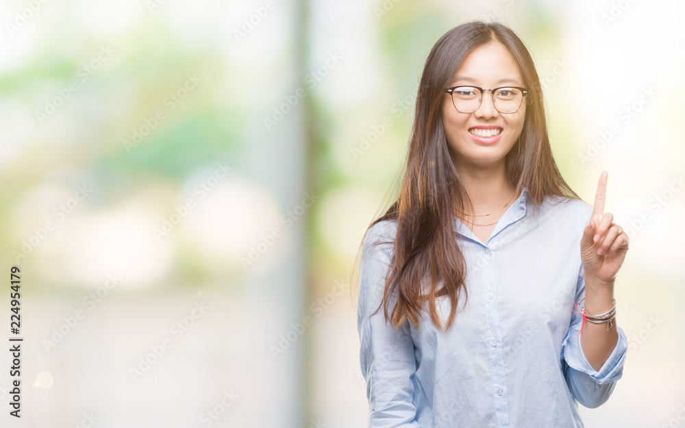 Young asian business woman wearing glasses over isolated background showing and pointing up with finger number one while smiling confident and happy. Young asian business woman wearing glasses over isolated background showing and pointing up with finger number one while smiling confident and happy.