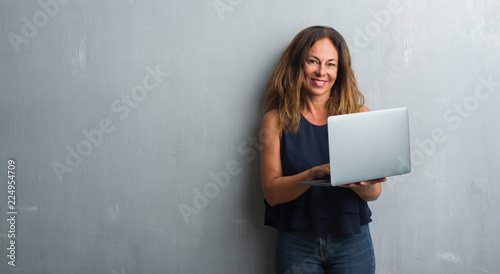 Middle age hispanic woman standing over grey grunge wall using laptop with a happy face standing and smiling with a confident smile showing teeth