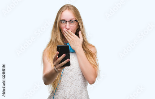 Blonde teenager woman holding smartphone and wearing headphones cover mouth with hand shocked with shame for mistake, expression of fear, scared in silence, secret concept