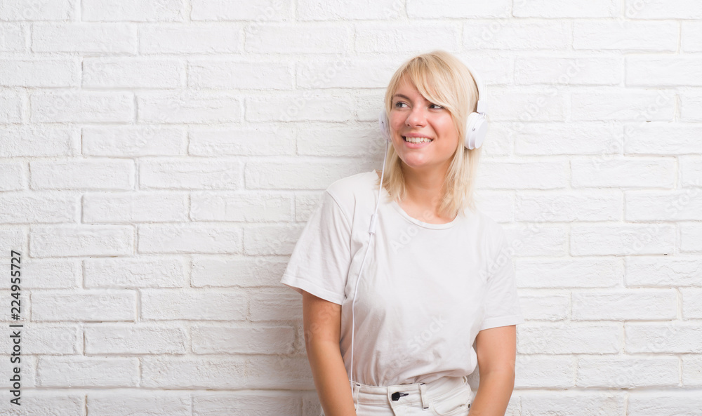 Adult caucasian woman over white brick wall wearing headphones looking away to side with smile on face, natural expression. Laughing confident. Adult caucasian woman over white brick wall wearing headphones looking away to side with smile on face, natural expression. Laughing confident.