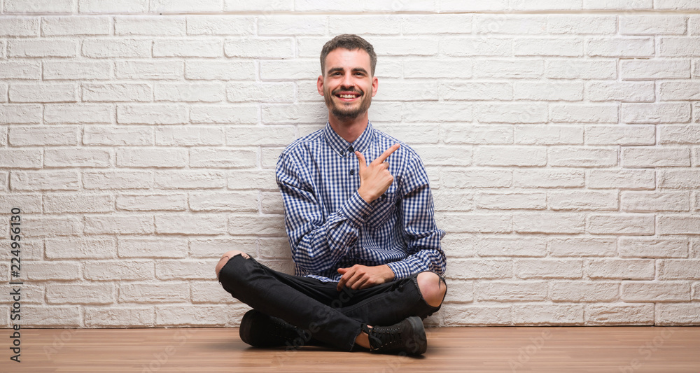 Young adult man sitting over white brick wall cheerful with a smile of face pointing with hand and finger up to the side with happy and natural expression on face looking at the camera. Young adult man sitting over white brick wall cheerful with a smile of face pointing with hand and finger up to the side with happy and natural expression on face looking at the camera.