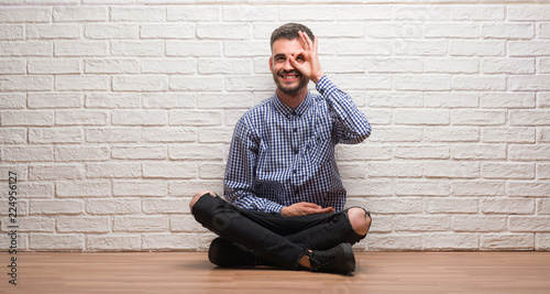 Young adult man sitting over white brick wall doing ok gesture with hand smiling, eye looking through fingers with happy face.