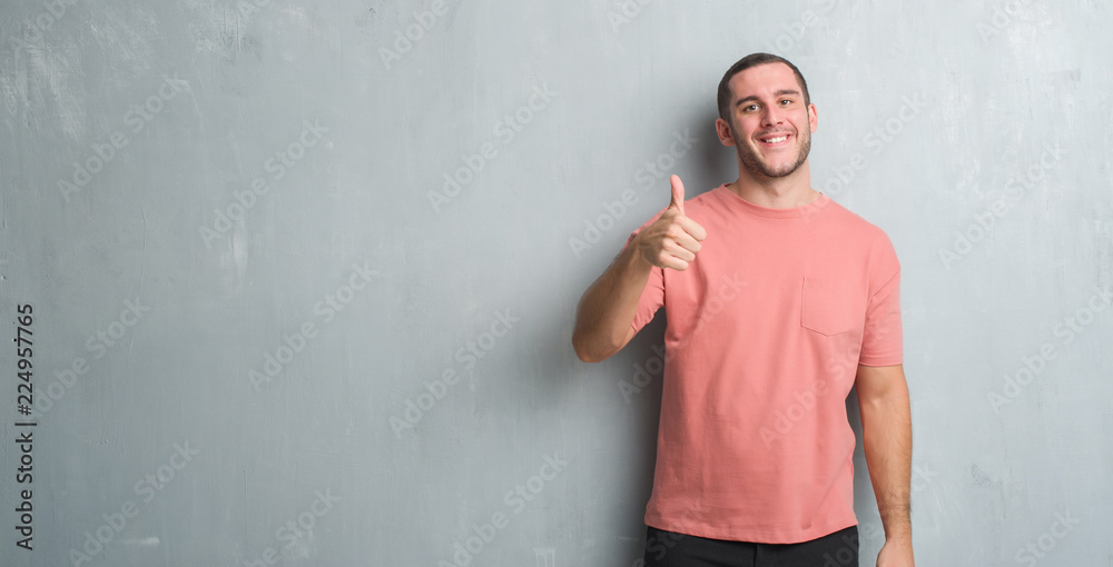 Young caucasian man over grey grunge wall doing happy thumbs up gesture with hand. Approving expression looking at the camera with showing success. Young caucasian man over grey grunge wall doing happy thumbs up gesture with hand. Approving expression looking at the camera with showing success.
