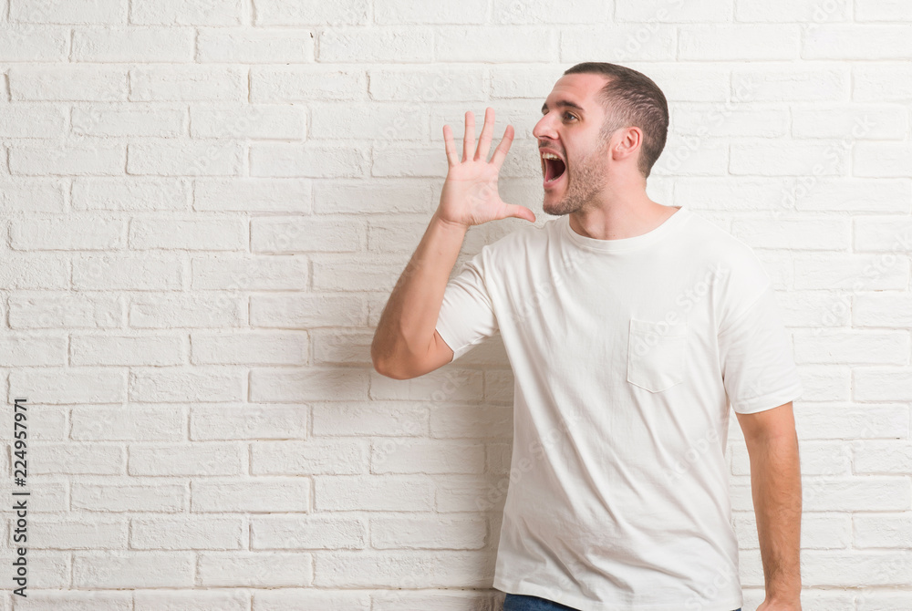 Young caucasian man standing over white brick wall shouting and screaming loud to side with hand on mouth. Communication concept. Young caucasian man standing over white brick wall shouting and screaming loud to side with hand on mouth. Communication concept.