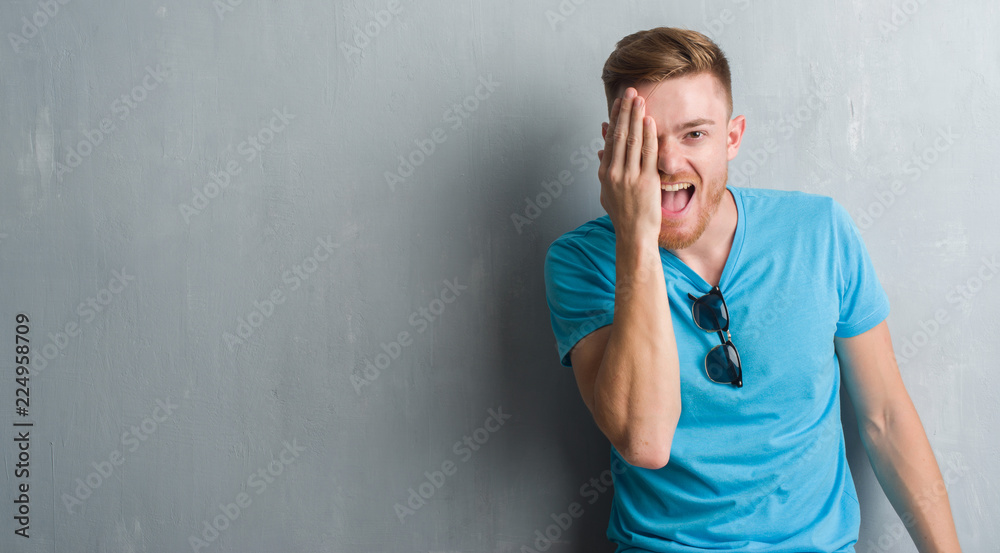 Young redhead man over grey grunge wall wearing casual outfit covering one eye with hand with confident smile on face and surprise emotion. Young redhead man over grey grunge wall wearing casual outfit covering one eye with hand with confident smile on face and surprise emotion.