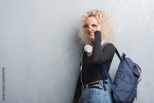 Young blonde woman over grunge grey wall wearing backpack and headphones doing ok gesture with hand smiling, eye looking through fingers with happy face.