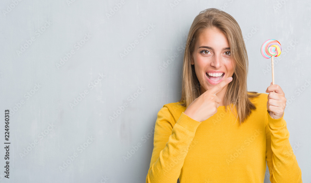 Beautiful young woman over grunge grey wall eating lollipop candy very happy pointing with hand and finger Beautiful young woman over grunge grey wall eating lollipop candy very happy pointing with hand and finger