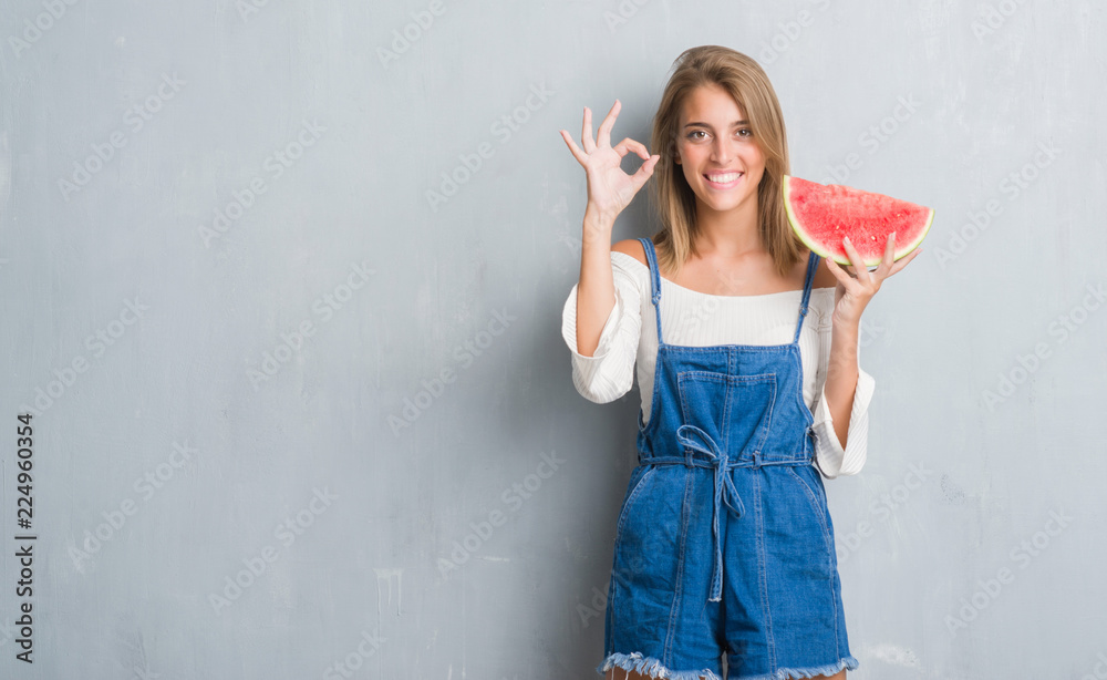 Beautiful young woman over grunge grey wall eating water melon doing ok sign with fingers, excellent symbol Beautiful young woman over grunge grey wall eating water melon doing ok sign with fingers, excellent symbol