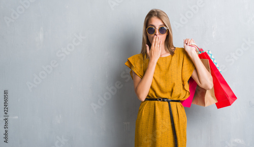 Beautiful young woman over grunge grey wall holding shopping bags on sales cover mouth with hand shocked with shame for mistake, expression of fear, scared in silence, secret concept