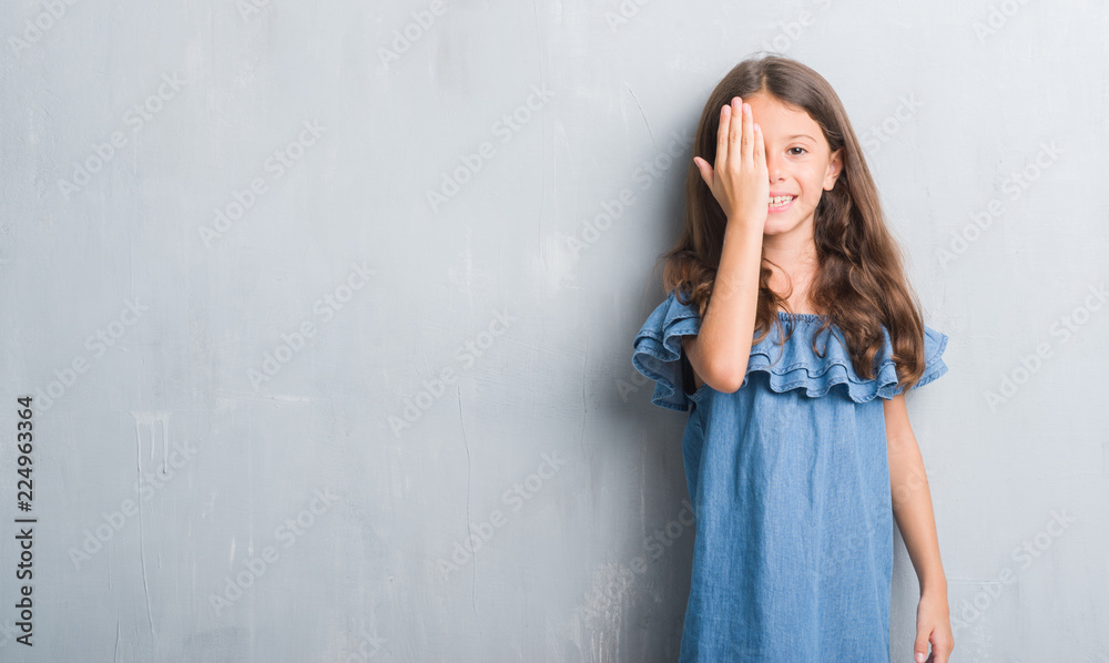 Young hispanic kid over grunge grey wall covering one eye with hand with confident smile on face and surprise emotion. Young hispanic kid over grunge grey wall covering one eye with hand with confident smile on face and surprise emotion.