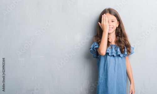 Young hispanic kid over grunge grey wall covering one eye with hand with confident smile on face and surprise emotion.