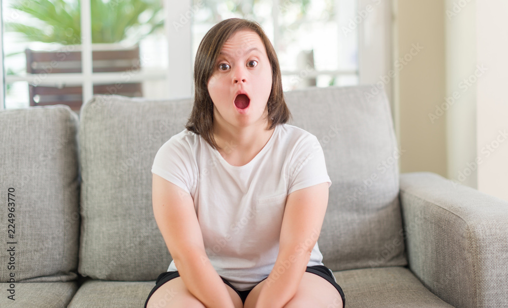 Down syndrome woman sitting on the sofa at home scared in shock with a surprise face, afraid and excited with fear expression Down syndrome woman sitting on the sofa at home scared in shock with a surprise face, afraid and excited with fear expression
