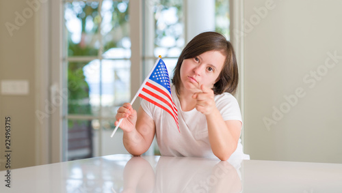 Down syndrome woman at home holding flag of usa pointing with finger to the camera and to you, hand sign, positive and confident gesture from the front