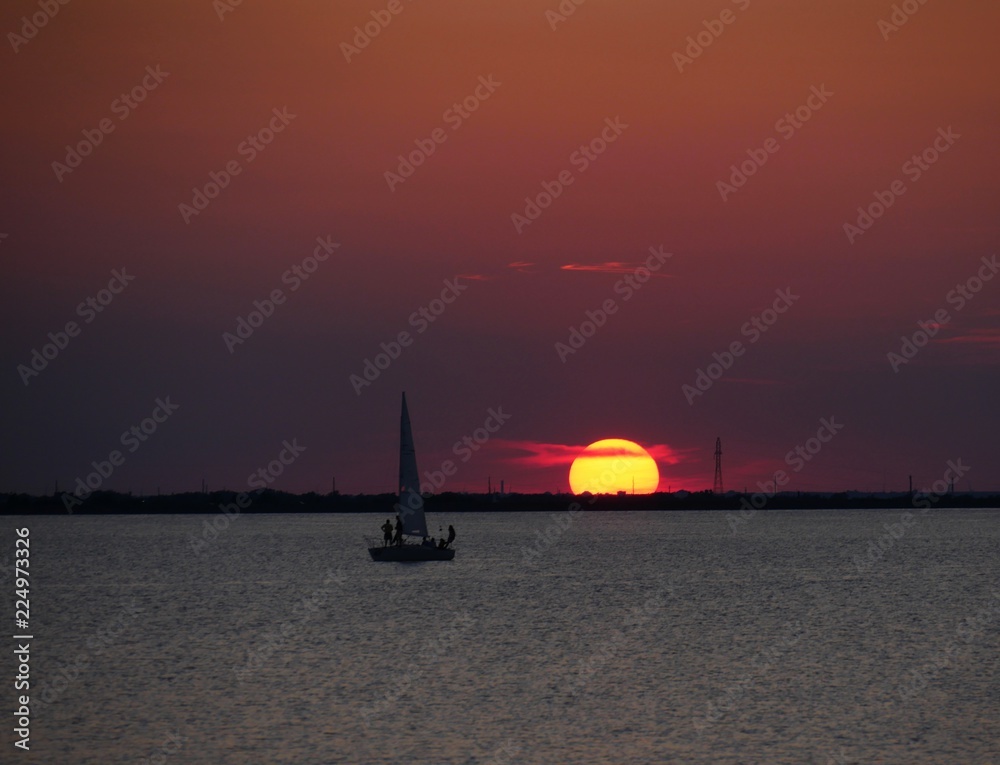 The sun starting to set in the horizon,  with a boat sailing some distance from it in the water