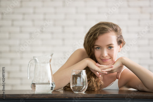 Beautiful young woman in joyful postures with jug and glass of drinking water on the side