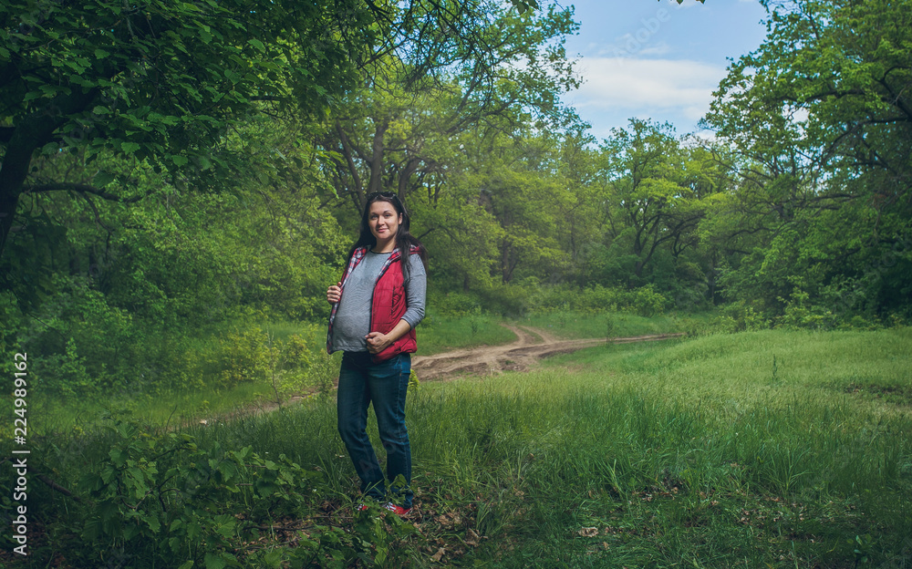Portrait of a young beautiful pregnant brunette girl who walks through the green forest, wearing jeans and a red vest. Portrait of a young beautiful pregnant brunette girl who walks through the green forest, wearing jeans and a red vest.