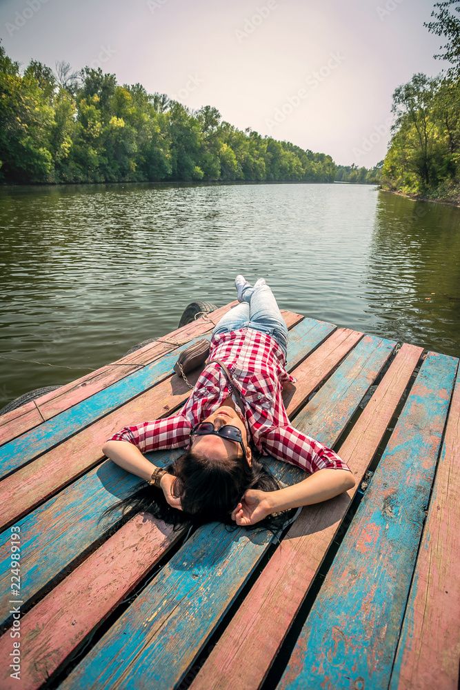 Young beautiful European girl basking in the sun lying on a river pontoon on a green forest background, wearing jeans and a plaid shirt Young beautiful European girl basking in the sun lying on a river pontoon on a green forest background, wearing jeans and a plaid shirt