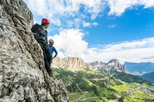 two young mountain climbers on a steep and exposed climb in the Dolomites of Alta Badia in Italy with a great view of the Val Gardena behind them
