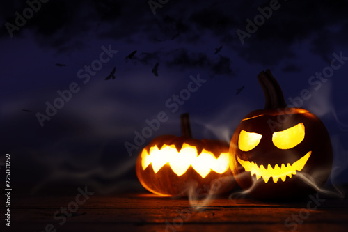 Halloween Dekoration auf Holz Untergrund, geschnitzte Kürbisse mit grusligen Fratzen und Lichtschein vor dunklem Nachthimmel