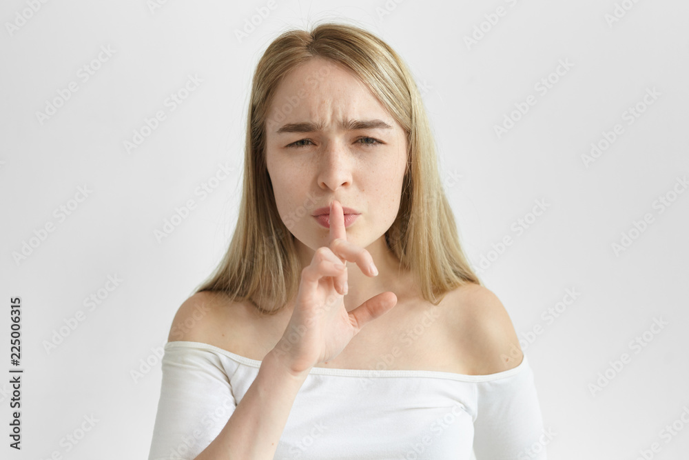 Body language. Attractive European girl in stylish top holding fore finger at her lips and frowning, asking to keep silent. Stylish young blonde female making hush gesture, saying Hush, be quiet Body language. Attractive European girl in stylish top holding fore finger at her lips and frowning, asking to keep silent. Stylish young blonde female making hush gesture, saying Hush, be quiet